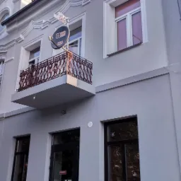 Odnowiona elewacja budynku z ozdobnymi detalami architektonicznymi, balkonem z kutą balustradą i anteną satelitarną na tle błękitnego nieba.