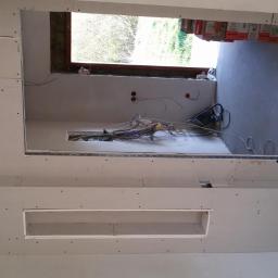 L&L Profi - Montaż płyt G-K na ścianie z widoczną instalacją elektryczną i otworami okiennymi. W tle worki z materiałami budowlanymi.