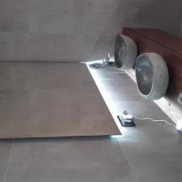 Nowoczesna łazienka z szarą, kwadratową glazurą, podświetlanym lustrem i umywalkami w kształcie kamiennych mis osadzonymi na drewnianej szafce.