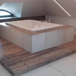 Nowoczesna wanna w obudowie z płytek, umieszczona na podwyższeniu z drewnopodobnych paneli w łazience na poddaszu z oknem dachowym.