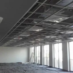 Wnętrze biura w trakcie remontu: widoczna konstrukcja sufitu podwieszanego z płyt kartonowo-gipsowych, częściowo wykończona ściana z fragmentem płyty w kolorze zielonym oraz rozłożona folia...