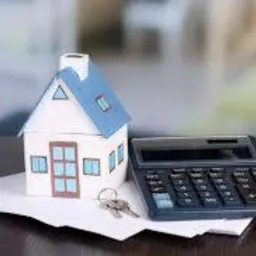 Model domu z niebieskim dachem, kalkulator i klucze leżące na białych kartkach, na ciemnym blacie stołu; w tle rozmyte, jasne pomieszczenie.