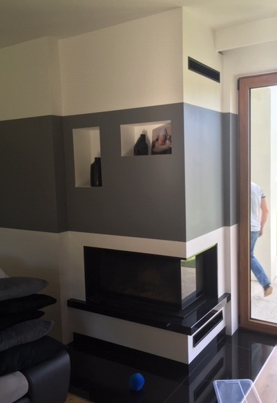 Nowoczesny narożny kominek z czarnym wkładem, wkomponowany w ścianę pomalowaną w poziome pasy szarości i bieli, z wnękami na dekoracje. Na podłodze błyszczące, czarne płytki. W tle widoczny...