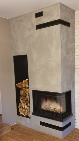 Nowoczesny kominek narożny z czarnymi akcentami i miejscem na drewno obok paleniska, wykończony szarą, teksturowaną ścianą.