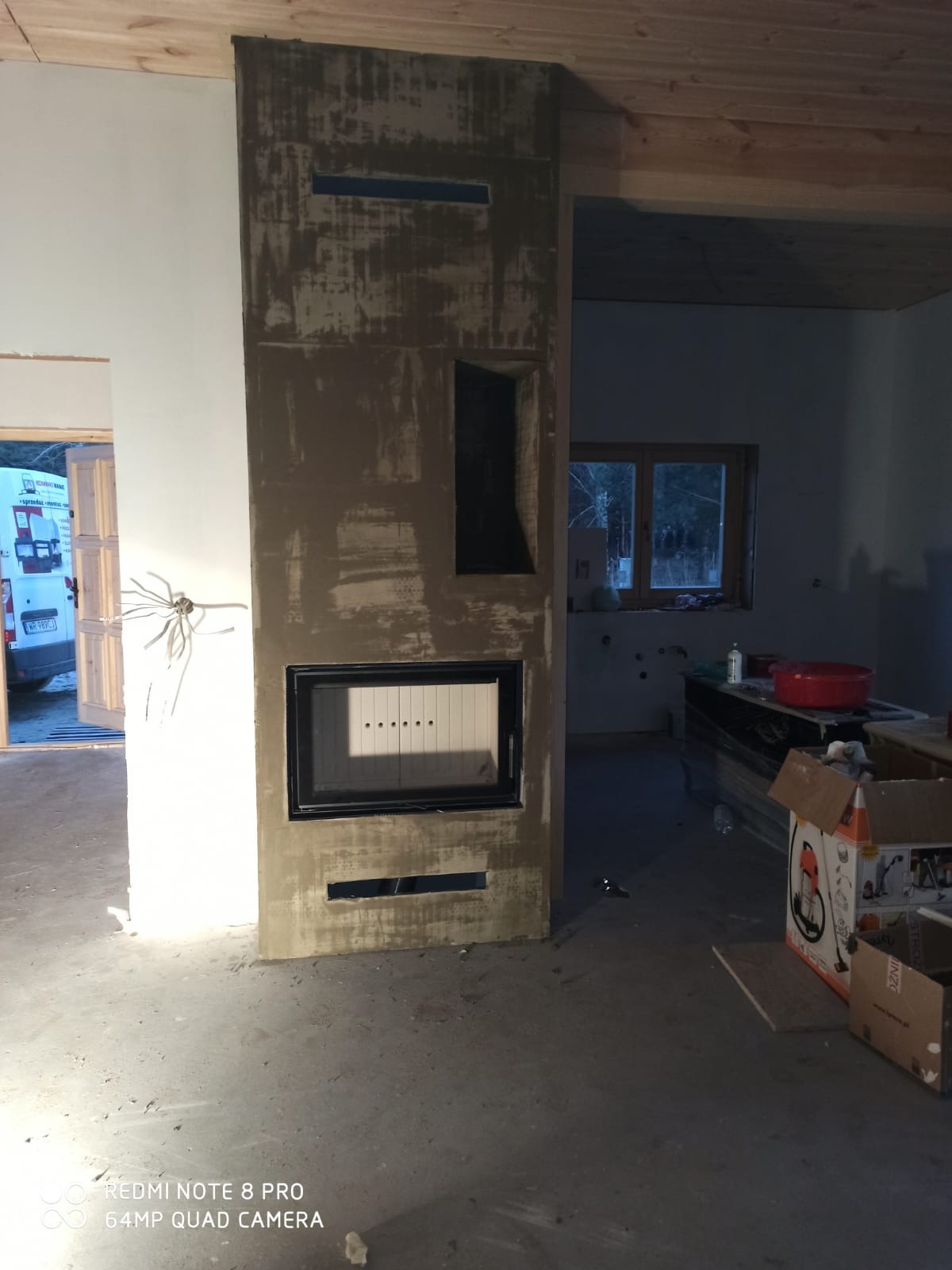 Wnętrze w trakcie wykańczania z widocznym kominkiem obłożonym surowym tynkiem, z otworami wentylacyjnymi i czarnym wkładem kominkowym, podłoga w stanie surowym, w tle okno i elementy wyposażenia.