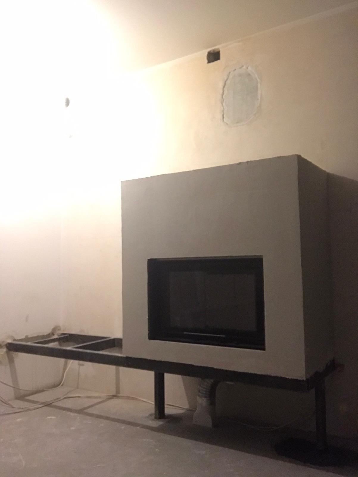 Surowy stan budowy kominka z czarnym wkładem, widoczna stalowa konstrukcja nośna i niedoskonałości na ścianie w tle.