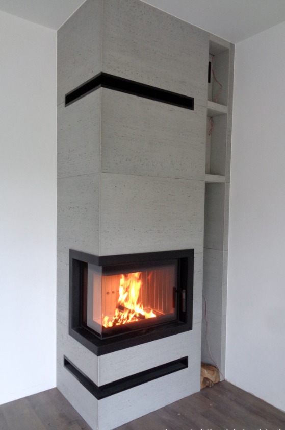 Nowoczesny narożny kominek obłożony jasnoszarym kamieniem z czarnymi akcentami, płomienie widoczne za szybą, obok wnęki na drewno lub dekoracje.