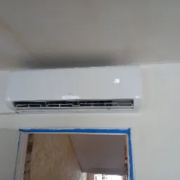 Biały klimatyzator ścienny marki AUX z wyświetlaną temperaturą 28 stopni, zamontowany nad ościeżnicą drzwi zabezpieczoną niebieską taśmą malarską.