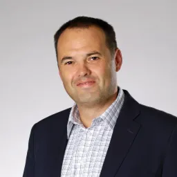 Portret mężczyzny w ciemnej marynarce i kraciastej koszuli na jasnoszarym tle. Uśmiechnięty, z krótkimi włosami.