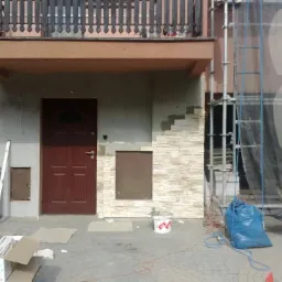 Fragment domu w trakcie prac elewacyjnych: częściowo wykończona kamienna okładzina, rusztowanie z siatką ochronną, narzędzia i materiały budowlane.