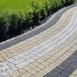 Falująca ścieżka z kostki brukowej w odcieniach beżu, bieli i szarości, obrzeże z ciemnej kostki, trawnik z niskimi krzewami w tle.