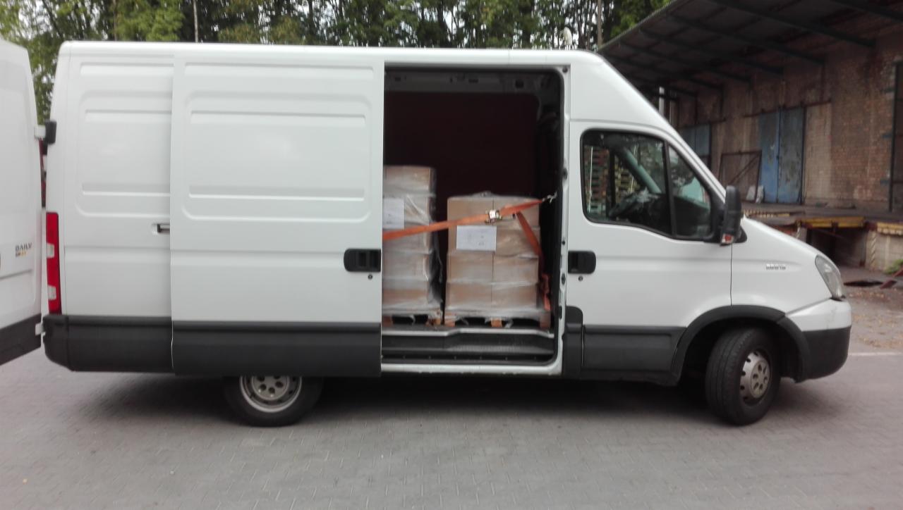 Biały bus z otwartymi drzwiami bocznymi, wewnątrz zabezpieczone pasami transportowymi kartony na paletach, widok z boku na tle budynku magazynowego.