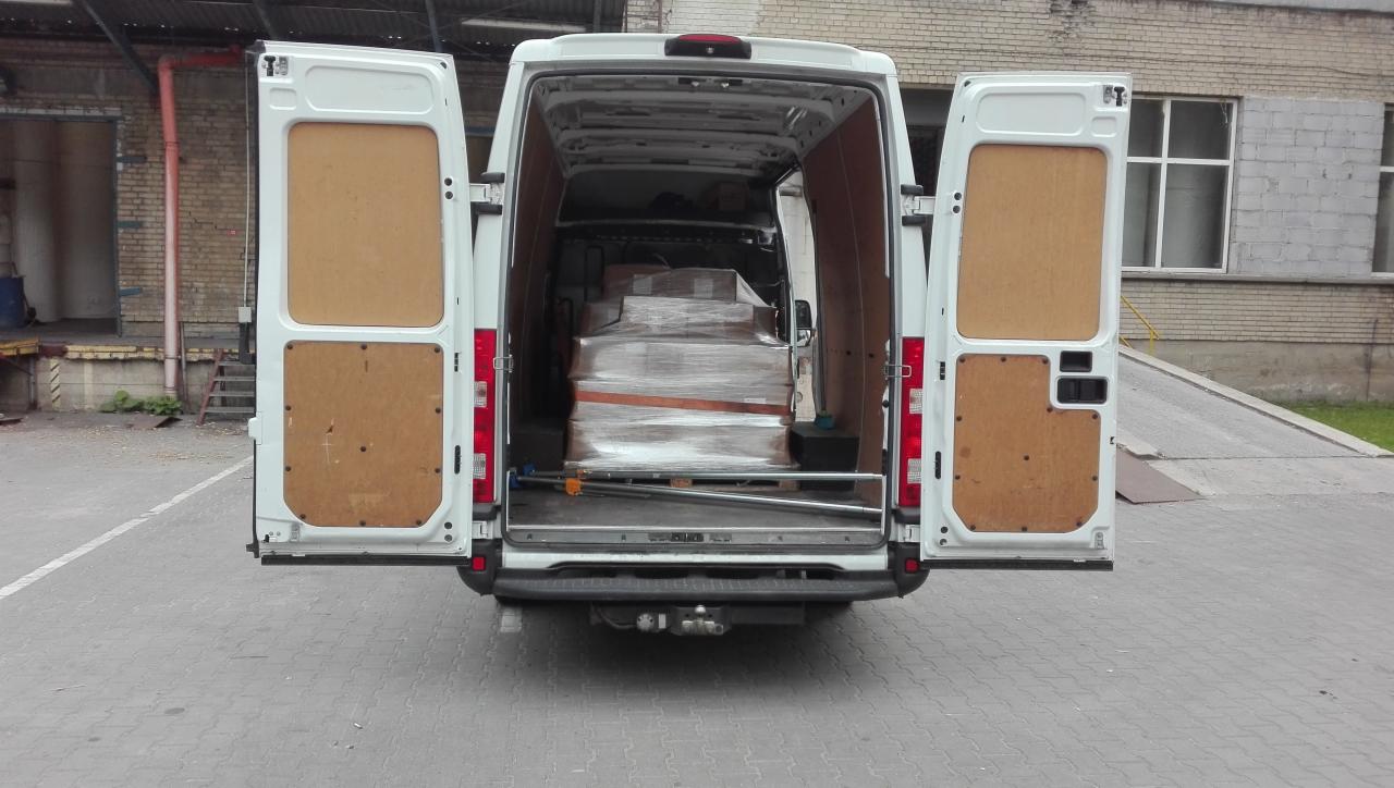 Widok z tyłu białego busa z otwartymi drzwiami, załadowanego kartonami owiniętymi folią na palecie, widoczny fragment rampy załadunkowej w tle.