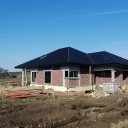 Budowa domu jednorodzinnego w stanie surowym otwartym, widoczne ściany z bloczków, czarny dach, okna bez szyb, teren wokół budynku z wykopami i materiałami budowlanymi.