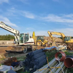Przygotowanie terenu pod budowę: trzy koparki (New Holland, Komatsu) na placu budowy, widoczne rury kanalizacyjne, zbrojenie i ogrodzenie tymczasowe, w tle wykop pod fundamenty.