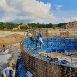 Budowa okrągłego zbiornika z szalunków i zbrojenia, widoczny żółty sprzęt budowlany w tle, pracownicy w trakcie montażu.
