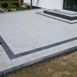 Wykończony taras z jasnoszarej kostki brukowej z ciemnoszarą obwódką i schodami, widoczny gumowy młotek i taczka, trawnik w tle.