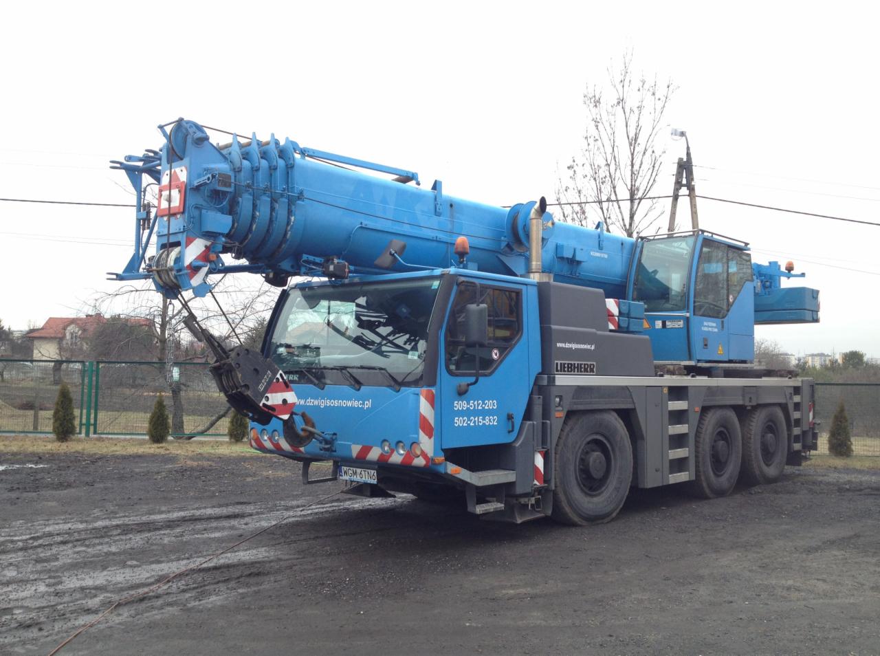 Niebieski dźwig Liebherr na błotnistym terenie; widoczne numery kontaktowe i adres strony www.dźwigisosnowiec.pl. Sprzęt budowlany w gotowości.