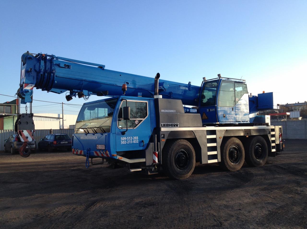 Niebieski dźwig Liebherr na placu budowy, widok z boku, z numerem telefonu i adresem strony internetowej na kabinie. Słońce oświetla maszynę z prawej strony.