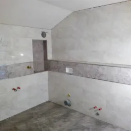 Wnętrze łazienki w trakcie remontu, widoczne ułożone jasne płytki z dekoracyjnym pasem, przygotowane instalacje wodne i kanalizacyjne, surowy mur przy wejściu.