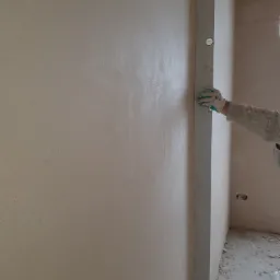 Świeżo nałożona, gładka warstwa tynku na ścianie, sprawdzana poziomicą przez pracownika w roboczych rękawicach; widoczne niedoskonałości podłoża przed dalszymi pracami wykończeniowymi.