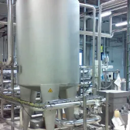 Błyszczący, stalowy zbiornik przemysłowy na trzech nogach, otoczony rurami, z żółtym znakiem ostrzegawczym na froncie, w hali produkcyjnej z kafelkowaną podłogą.