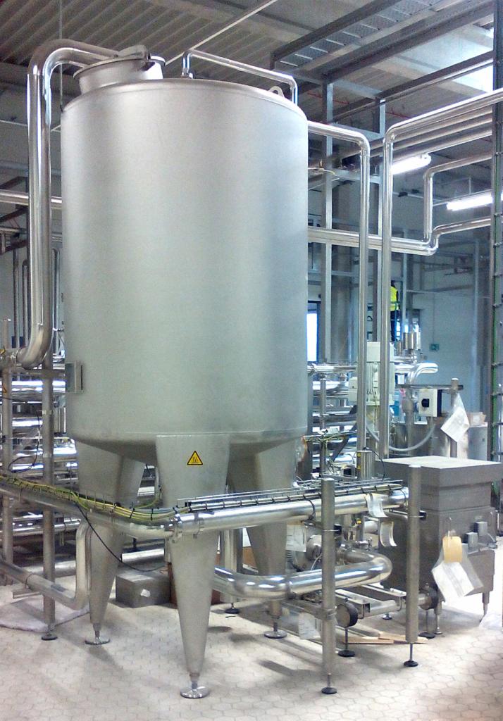 Błyszczący, stalowy zbiornik przemysłowy na trzech nogach, otoczony rurami, z żółtym znakiem ostrzegawczym na froncie, w hali produkcyjnej z kafelkowaną podłogą.