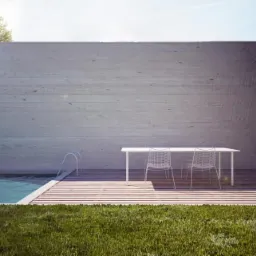 Minimalistyczny ogr&oacute;d z basenem, betonową ścianą i białym stołem z krzesłami na drewnianym tarasie, z widoczną drabinką do basenu i starannie przystrzyżonym trawnikiem.