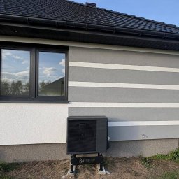 HYDROM Instalacje Grzewcze i Sanitarne - Czarna pompa ciepła Panasonic zamontowana na zewnątrz budynku z szarą elewacją w białe pasy. W tle widoczny firmowy bus i fragment krajobrazu.