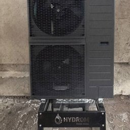 HYDROM Instalacje Grzewcze i Sanitarne - Tomasz Roman - Ciemnoszara jednostka zewnętrzna pompy ciepła Panasonic z dwoma wentylatorami, zamontowana na czarnym stojaku HYDROM na betonowych płytach.