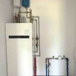 HYDROM Instalacje Grzewcze i Sanitarne - Tomasz Roman - Nowoczesna, biała pompa ciepła z dwoma zbiornikami wyrównawczymi (białym i niebieskim) oraz precyzyjnie poprowadzonymi instalacjami rurowymi w jasnym pomieszczeniu technicznym.