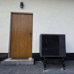 HYDROM Instalacje Grzewcze i Sanitarne - Tomasz Roman - Zainstalowana czarna pompa ciepła Panasonic na metalowym stojaku Hydrom, stojąca na ciemnej kostce brukowej obok drewnianych drzwi i jasnej ściany budynku z rynną. Widoczna skrzynka narzędziowa.