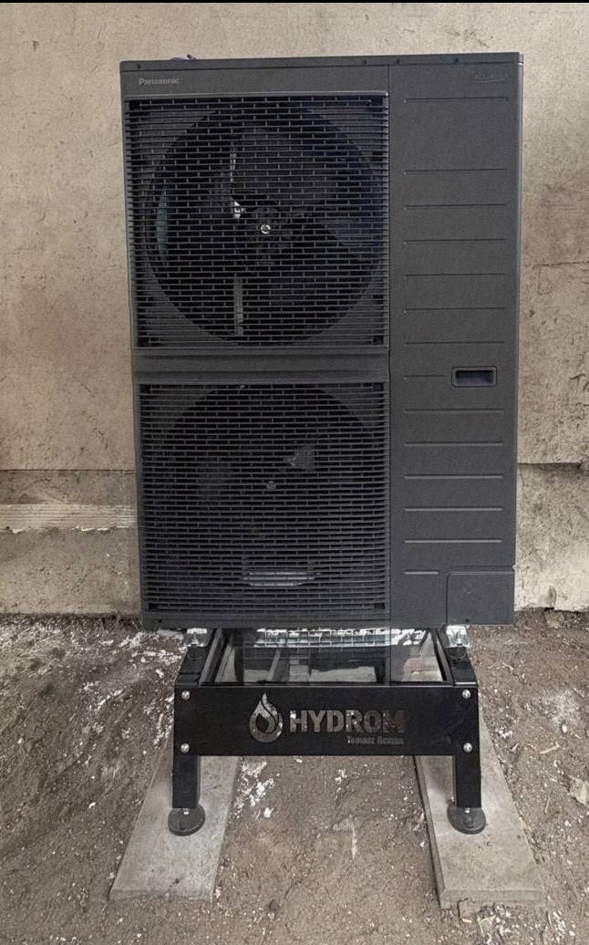 Czarna pompa ciepła Panasonic, ustawiona na metalowej podstawie z logo Hydrom, na betonowych płytach. Widoczne dwa wentylatory i szara ściana w tle.