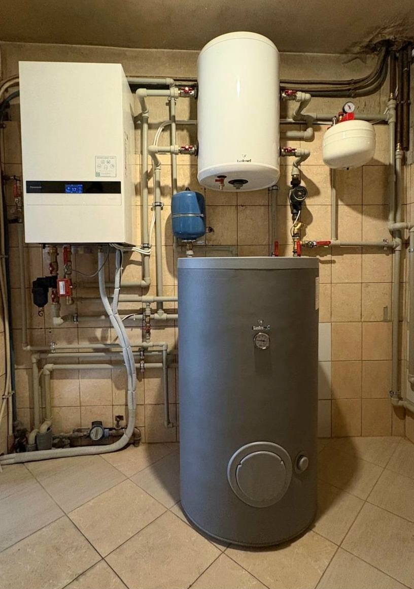 Pompa ciepła Panasonic z zbiornikami i rurami w kotłowni z beżowymi płytkami. Widoczne liczne przyłącza i zawory. Ujęcie z dołu, podkreślające wysokość instalacji.