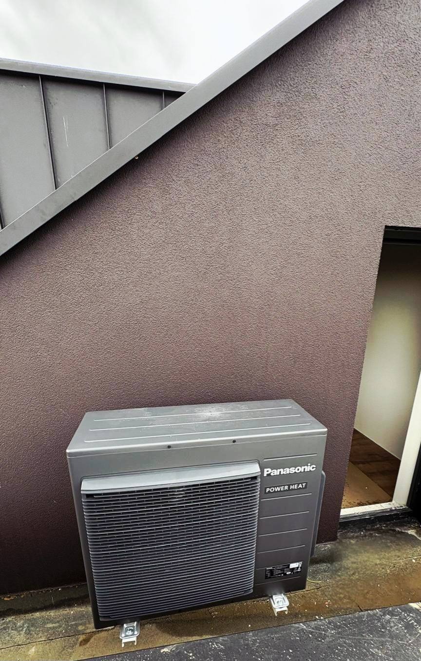 Szara, zewnętrzna jednostka klimatyzacji Panasonic Power Heat zamontowana przy brązowej ścianie budynku z widocznym fragmentem dachu i wejściem do pomieszczenia.