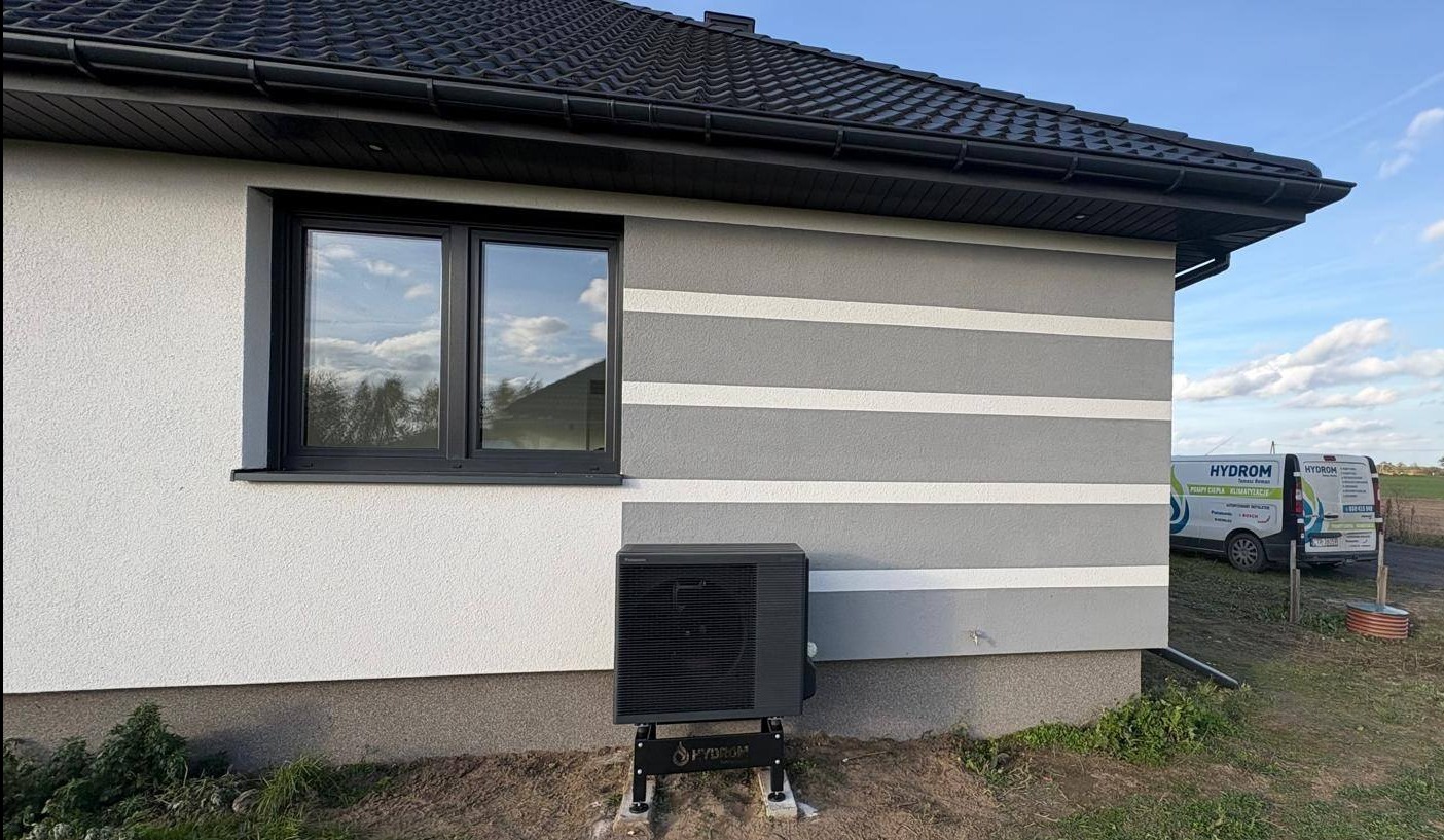 Czarna pompa ciepła Panasonic zamontowana na zewnątrz budynku z szarą elewacją w białe pasy. W tle widoczny firmowy bus i fragment krajobrazu.