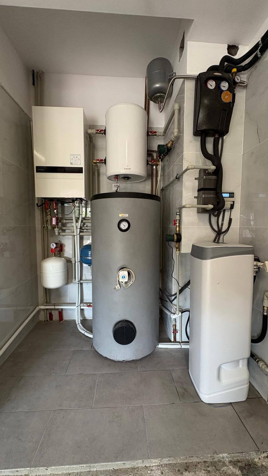 Zestaw urządzeń grzewczych w kotłowni: bojler, zbiornik wyrównawczy, piec gazowy kondensacyjny i pompa ciepła. Instalacja w pomieszczeniu wykończonym płytkami.