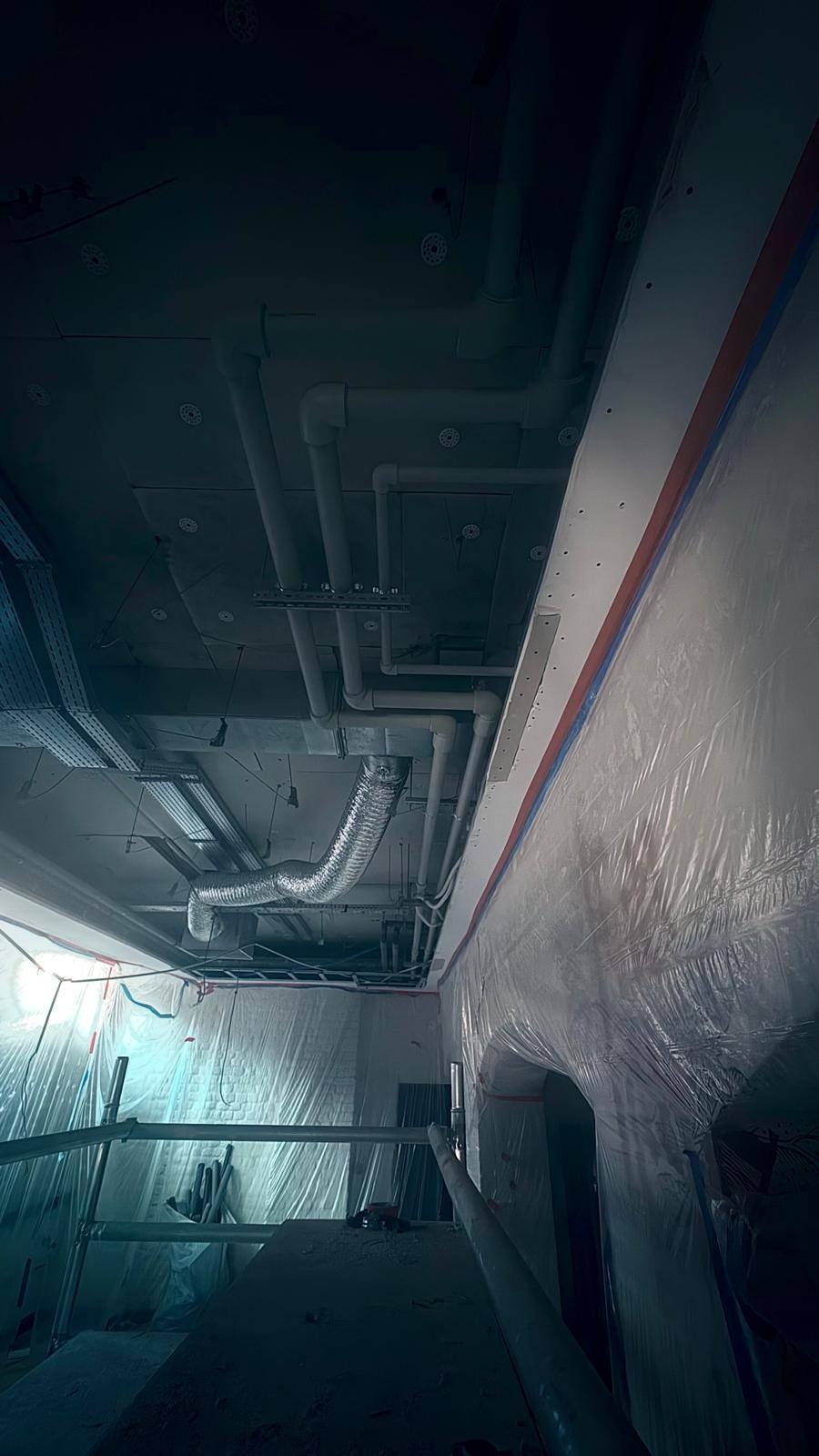 Instalacja rur wentylacyjnych i hydraulicznych pod sufitem w surowym wnętrzu, zabezpieczonym folią malarską. Widoczne kable i elementy konstrukcyjne.