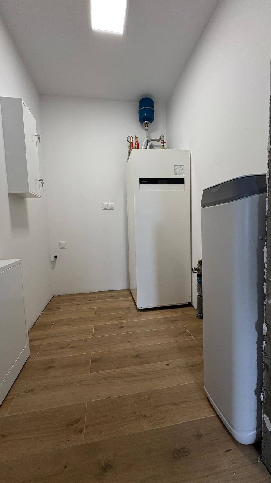 Biała pompa ciepła Panasonic z niebieskim zbiornikiem wyrównawczym w nowo wykończonym pomieszczeniu z drewnopodobną podłogą i białymi ścianami.