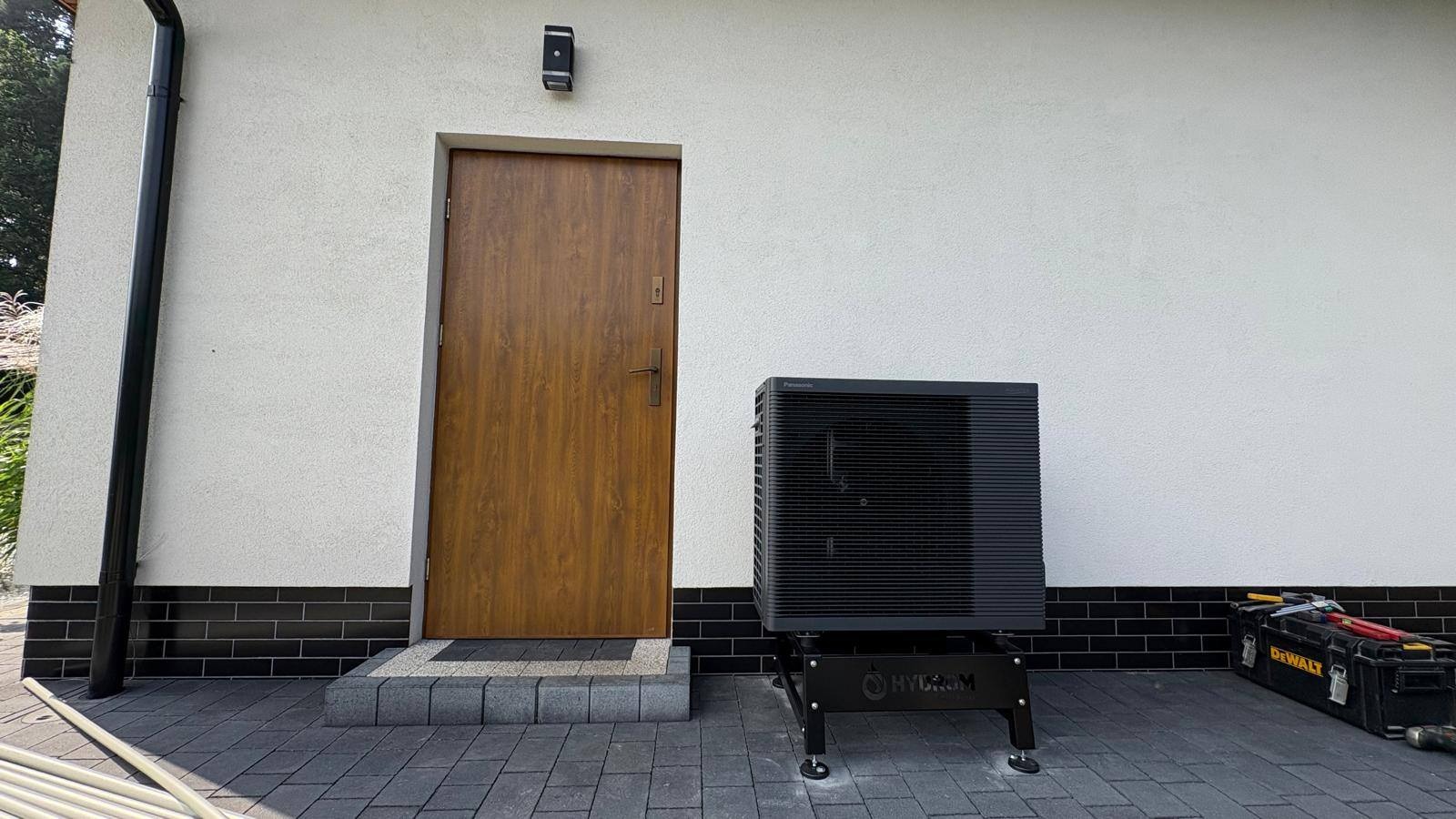 Czarna pompa ciepła Hydrom zamontowana przy ścianie domu z brązowymi drzwiami wejściowymi. Obok skrzynka z narzędziami DeWalt.