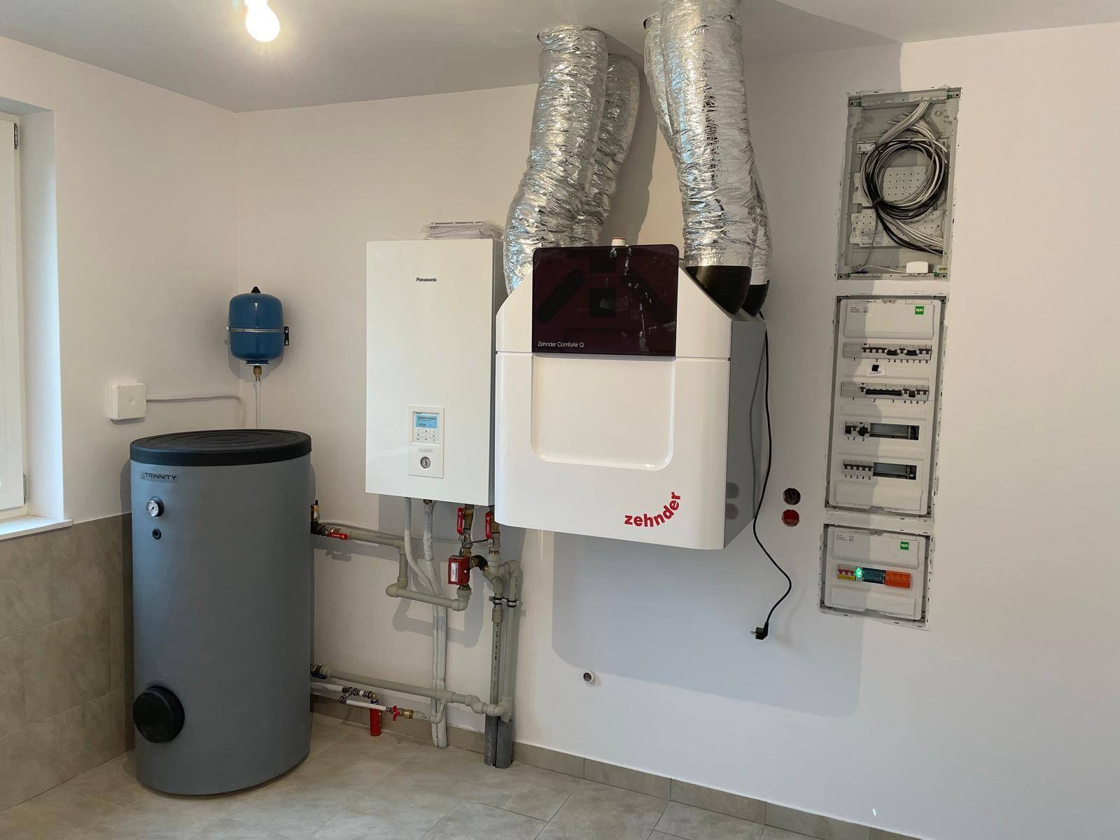 Zestaw urządzeń grzewczych: zbiornik Trinity, piec Panasonic, centrala Zehnder ComfoAir Q z przewodami wentylacyjnymi i rozdzielnia elektryczna na białej ścianie.