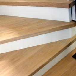 Wood & Iron Manufacture - Jasne, drewniane schody z białymi podstopnicami i czarną, ozdobną balustradą. Ujęcie z dołu, podkreślające detale wykończenia i wzór drewna.