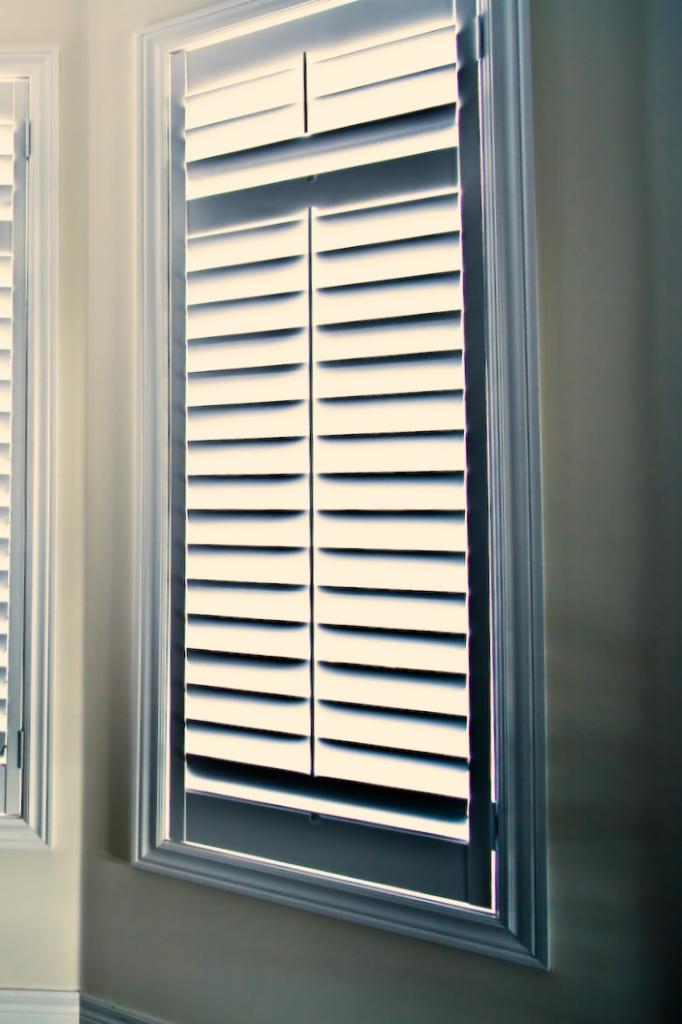 Zamknięte, białe żaluzje okienne w eleganckiej ramie, z widocznym fragmentem drugiej żaluzji po lewej stronie. Stonowana kolorystyka, subtelne cieniowanie.