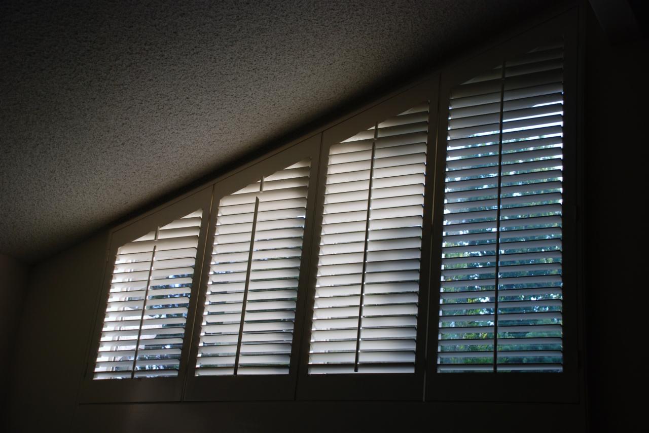 Trzy okna z białymi, drewnianymi żaluzjami w skośnej ścianie poddasza, przepuszczające rozproszone światło dzienne. Widok na zieleń za oknem.