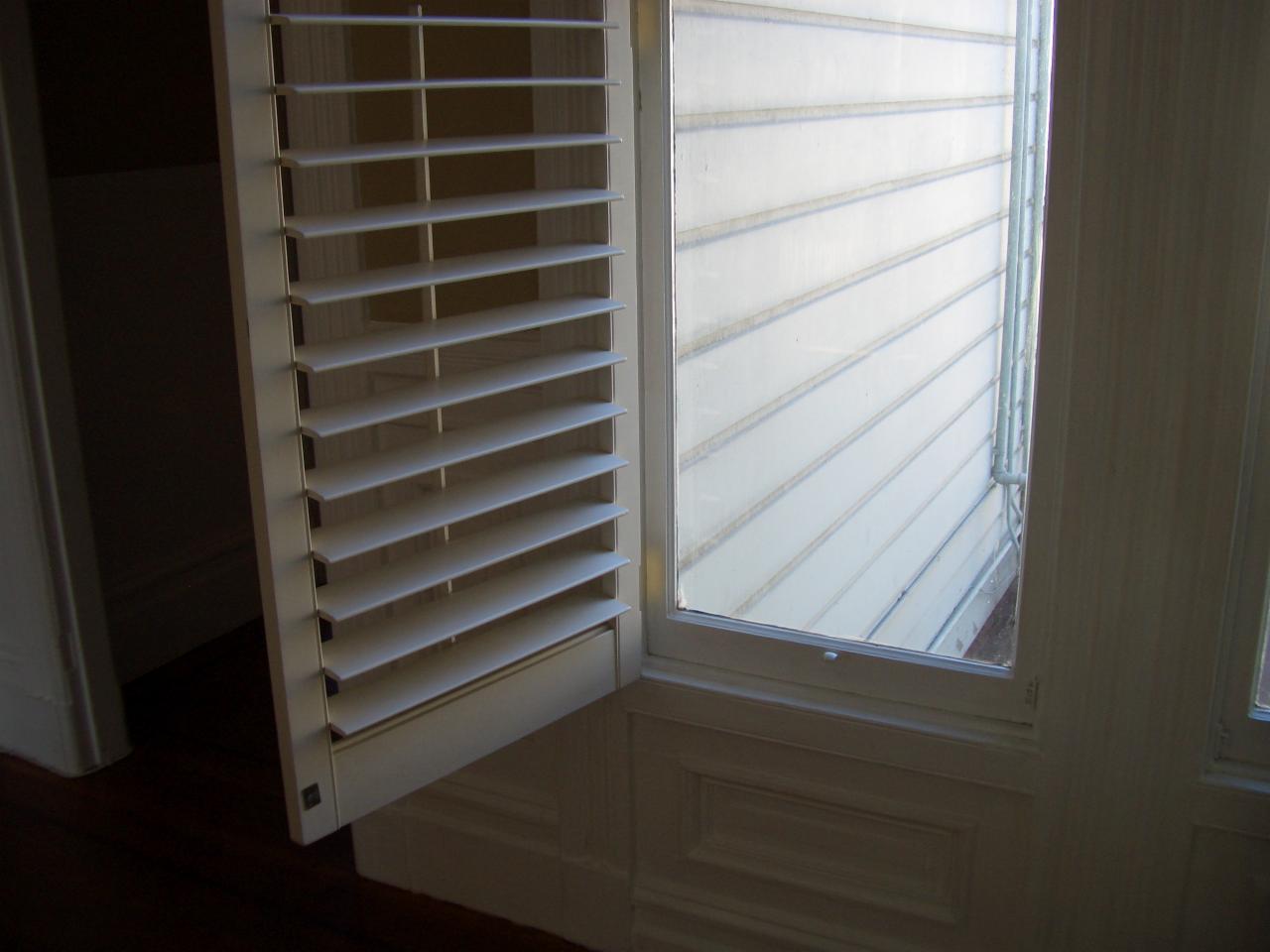 Białe okiennice żaluzjowe uchylone, odsłaniające widok na elewację budynku. Widoczne detale ramy okiennej i pionowe deski elewacyjne. Częściowo widoczna podłoga.