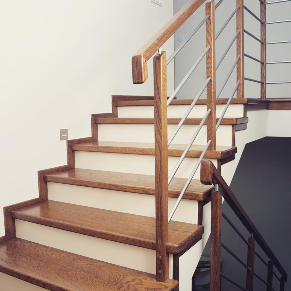 Nowoczesne schody drewniane z metalową balustradą. Stopnie w ciepłym odcieniu drewna kontrastują z białymi podstopnicami. Minimalistyczny design, jasne wnętrze.