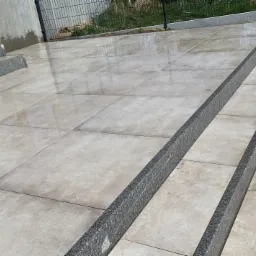 Wyłożony jasnymi, kwadratowymi płytami taras z granitowymi obrzeżami i schodami, widoczne mokre plamy na powierzchni płyt.