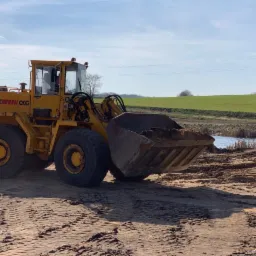Żółta ładowarka Kaellble CKG z łyżką pełną ziemi na terenie budowy, w tle zielone pole i małe oczko wodne.