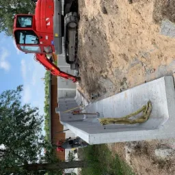 Czerwona koparka przy betonowym fundamencie w wykopie, widoczne żółte pasy mocujące, teren częściowo wyrównany.