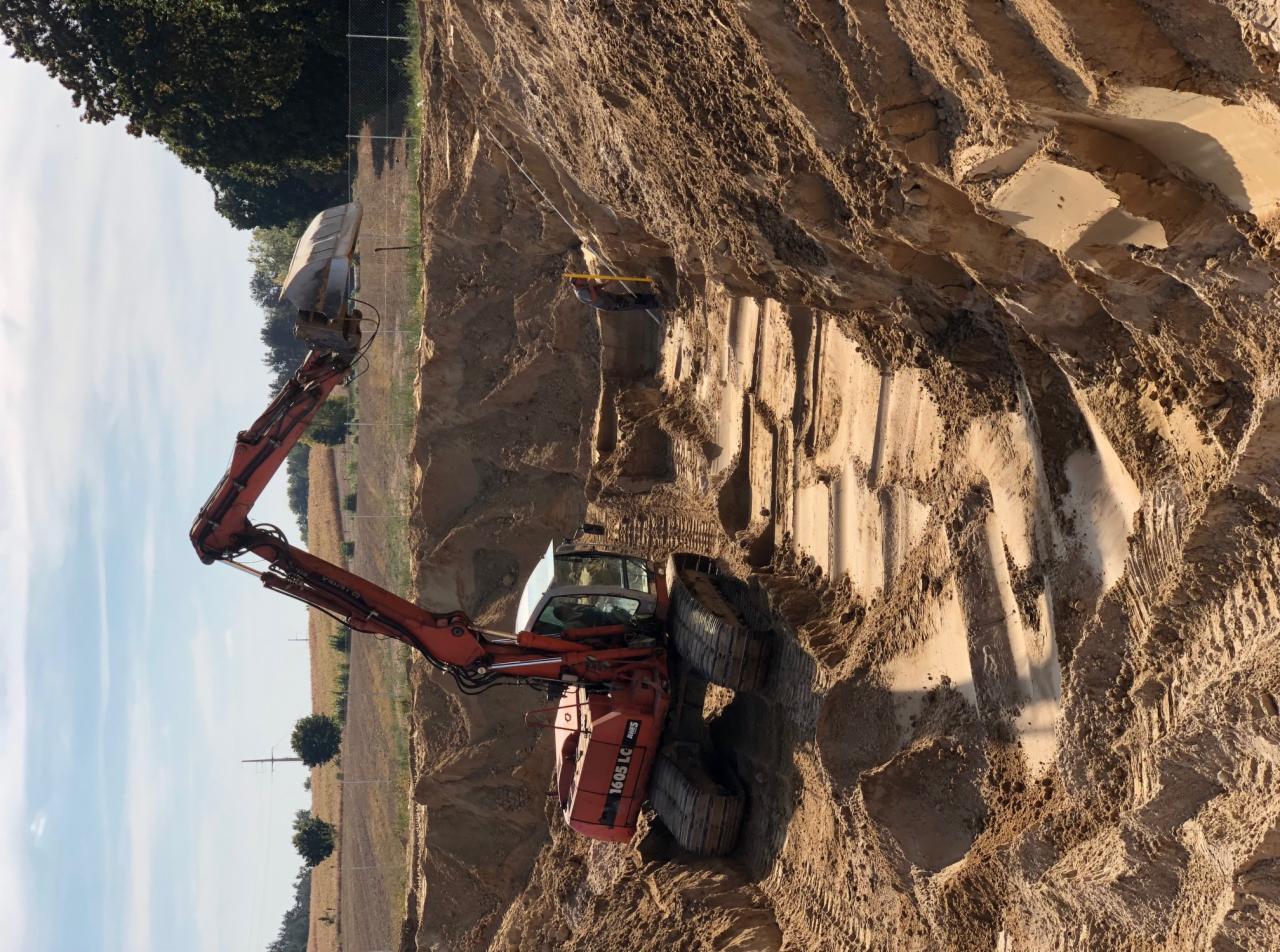 Czerwona koparka gąsienicowa wykonuje wykop w piaszczystej glebie, widoczne ślady gąsienic i wykopane rowy w ziemi.